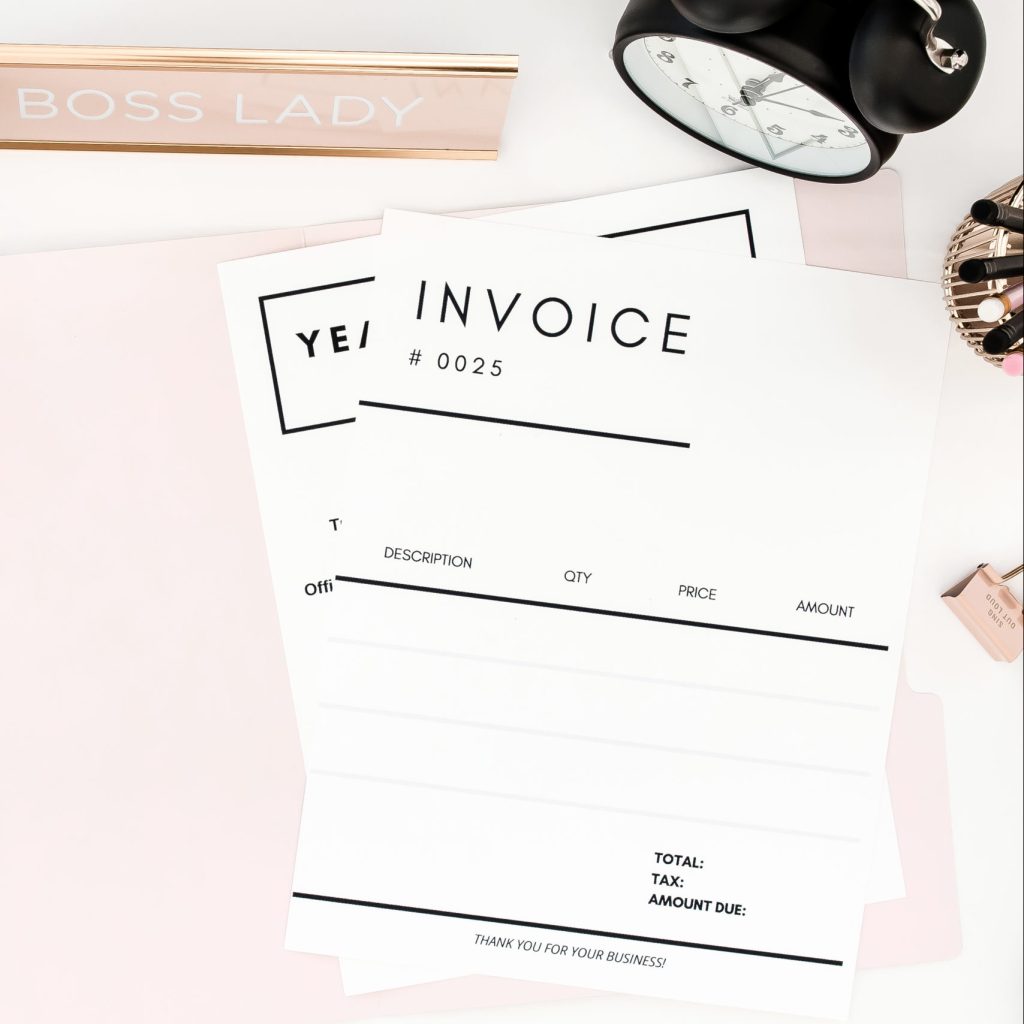 Two paper invoices, on a pink notepad with a boss lady desk sign and clock in the background