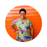 Keneena from Kablooie wearing a bright printed dress green glasses standing in front of a bright orange roller door