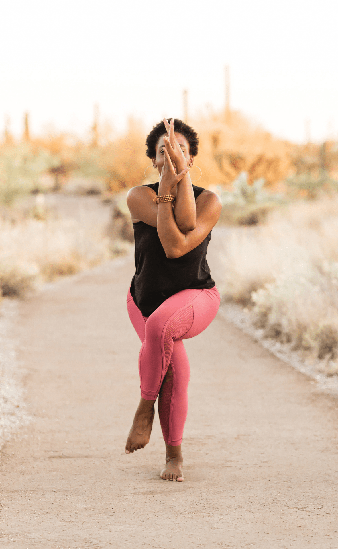 Black woman in black top pink pants doing garudasana or eagle yoga pose in a desert style background
