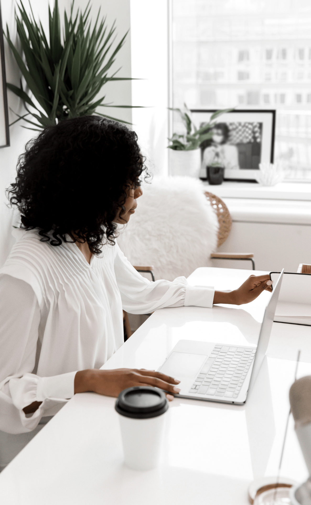 a woman of colour working at a white desktop with laptop and plants in the background