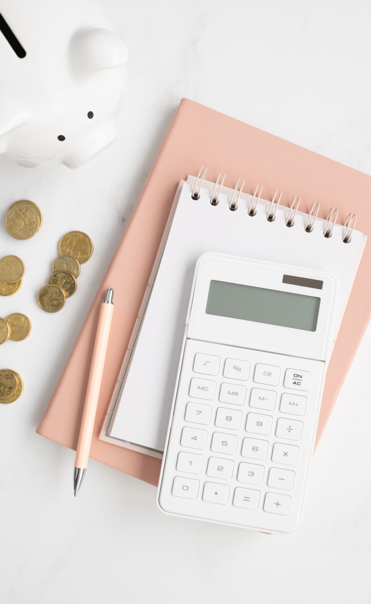 white desktop with peach notepad, white calculator, pens, gold coins and white piggy bank