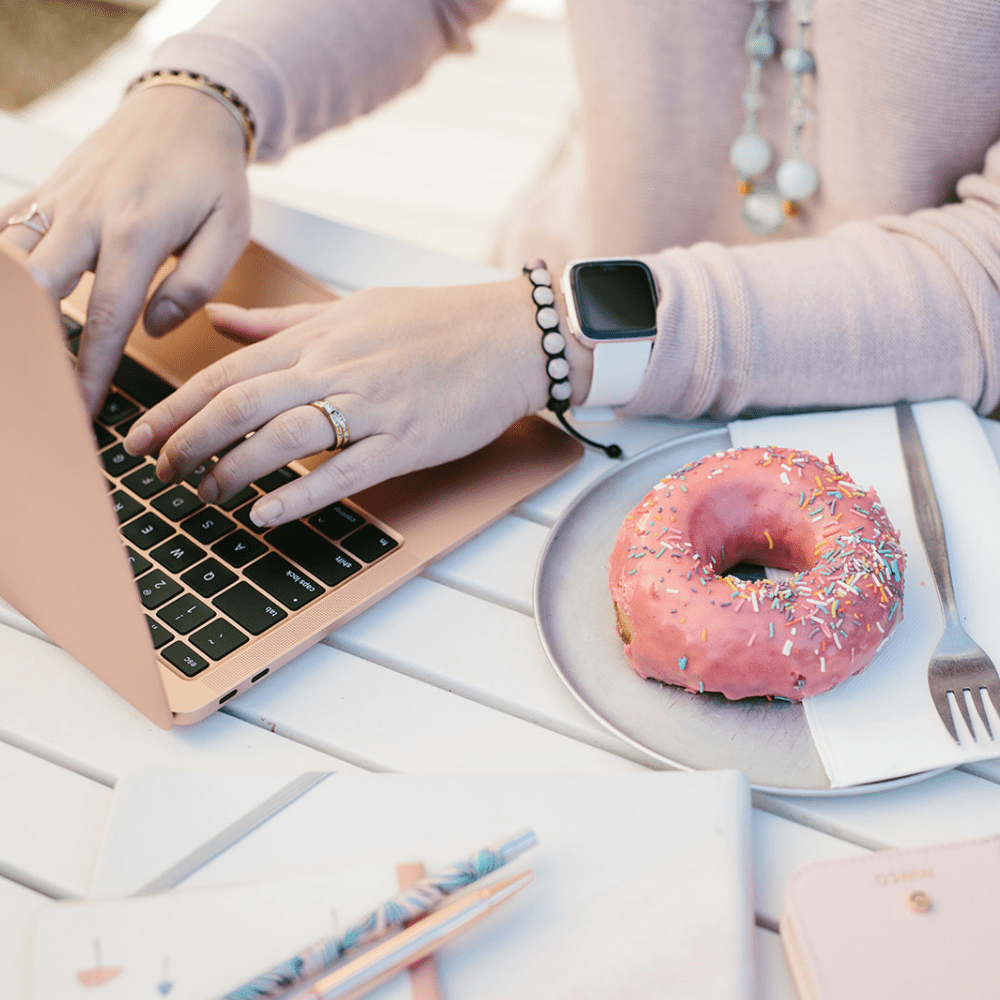 A shot of Lisa Turner's hands working at her bronze coloured laptop with a doughnut and cutlery to her side. Stationary and books to her front. Lisa is a Small Business Bookkeeper.