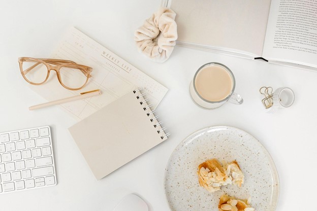 Neutral and peach toned flatlay desktop with a plate with pastry, coffee, scrunchie, glasses, notepads book and keyboard.