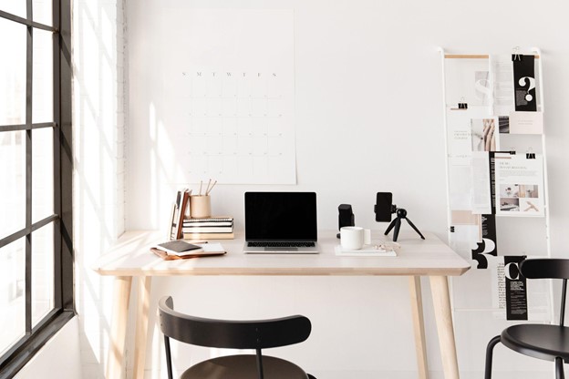 Neutral workspace with calendar on the wall above a wooden desk with pile of books, phone on tripod, coffee and laptop, with two black chairs.