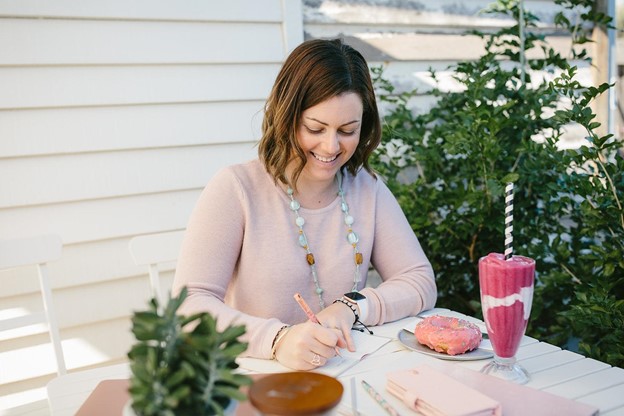 Lisa Turner seated at outdoor white table with pink donut and fruit smoothie writing in a notepad with other notebooks and phone on the table
