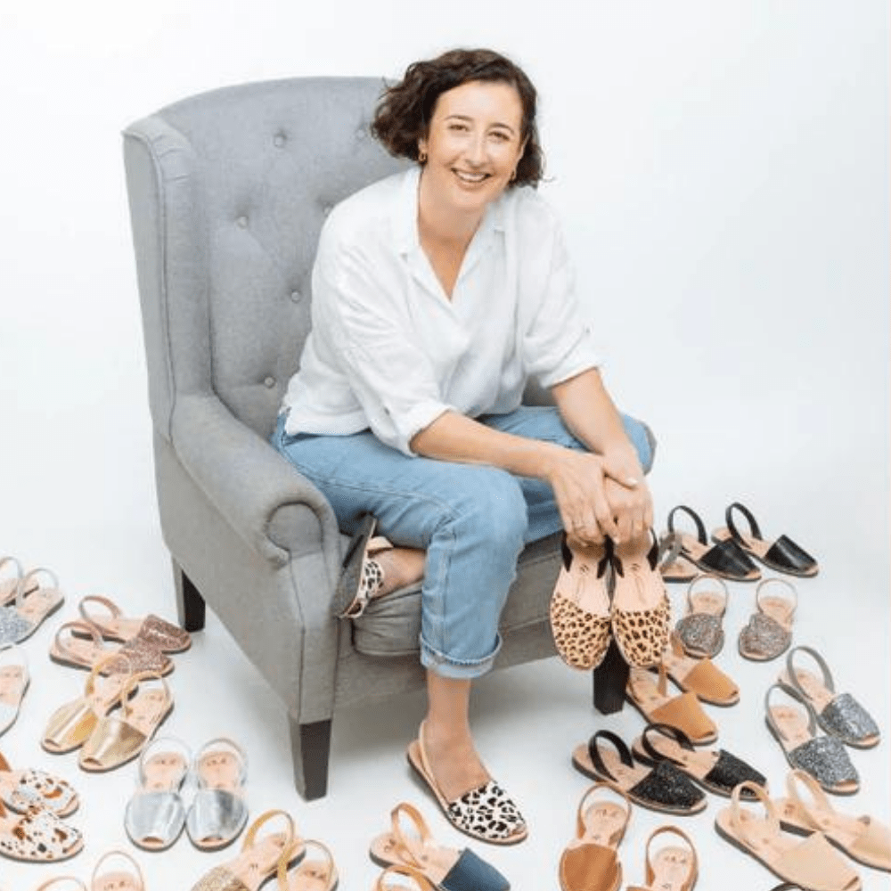 Naomi, a business owner seated on a large grey chair surrounded by many pairs of shoes, pictured as a client testimonial for the services of Accounted For You, a Bookkeeping service for women led business.