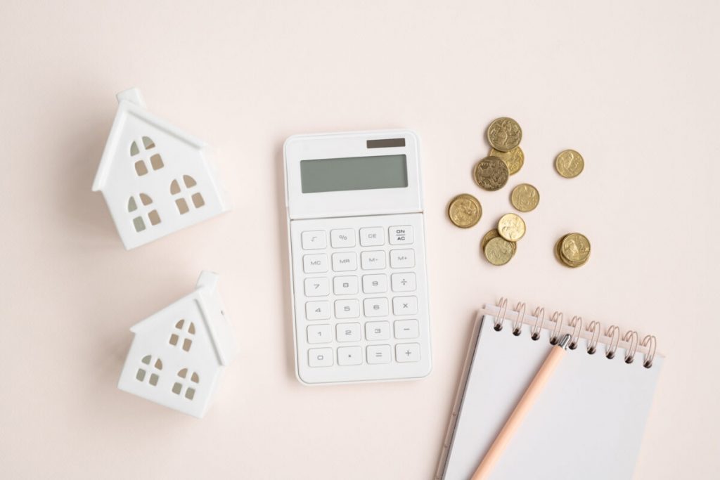 blush flatlay with two white house money boxes, white calculator, gold coins and notepad with pink pen.