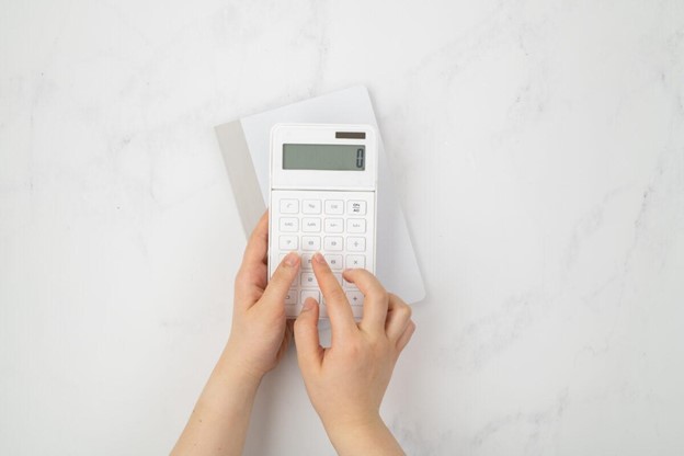 white marble desk flatlay with white calculator with notepaid behind it and childs hands typing on the numbers