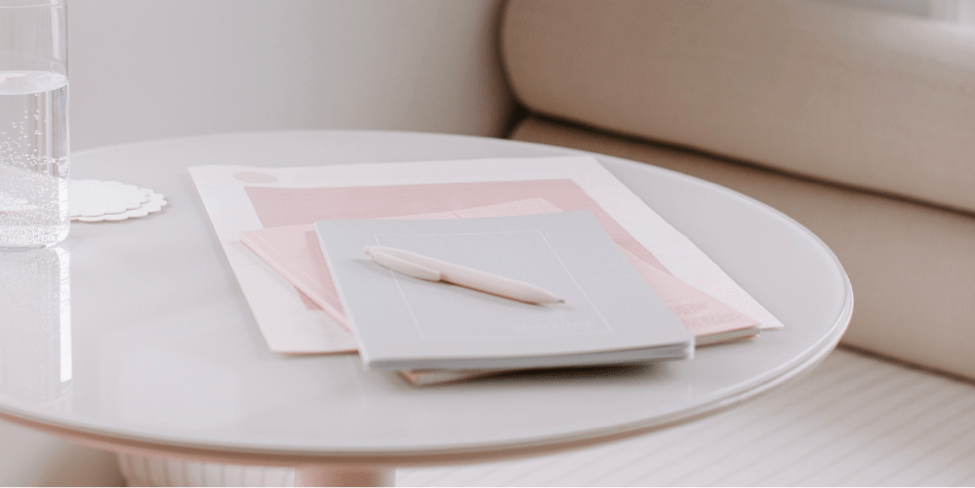 Pink notebooks and pen lying on a round coffee table beside a glass of water and near a beige lounge
