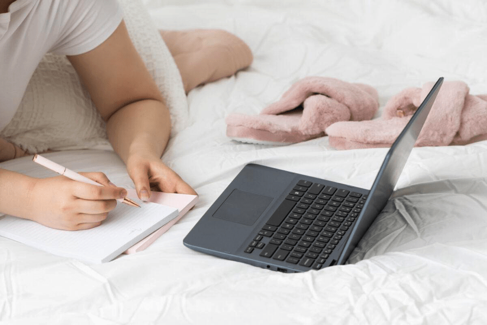 Girl lying on bed with notebook and pen in hand, with a laptop and slippers to the side.