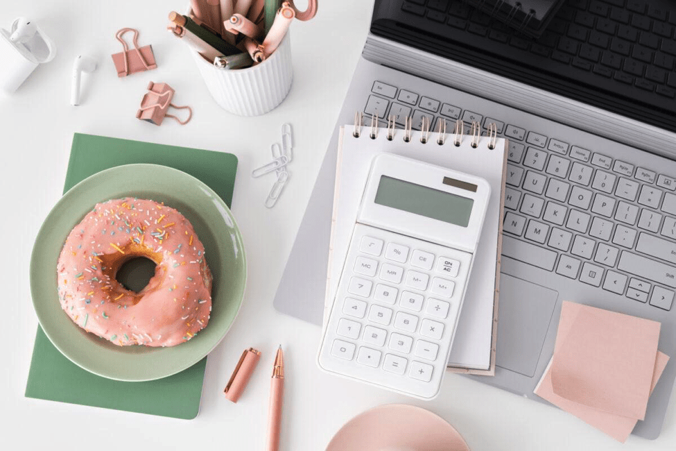 Desktop pictured from above with silver laptop open, notebook, calculator, pink post-it notes, pot of pens, green notebook and pink iced donut on a green plate with stationery accessories scattered on the desk.