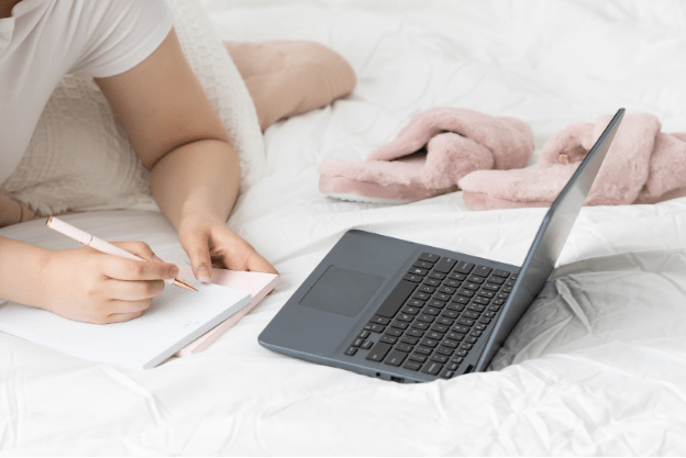 Woman writing in notebook on bed with white sheets with black laptop and pink slippers to the side