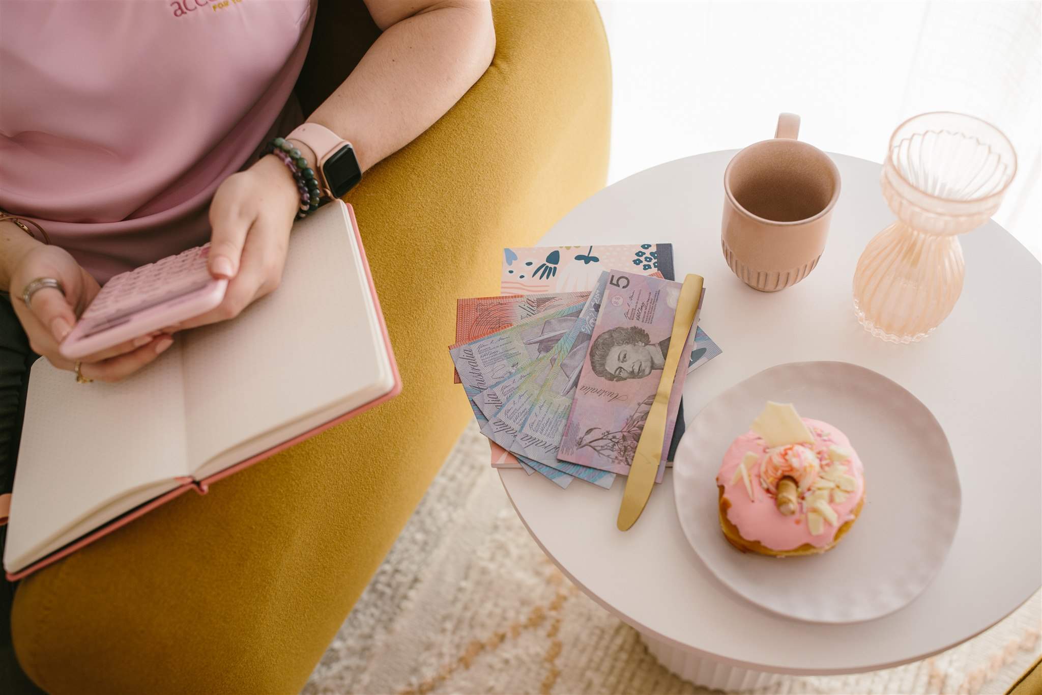 Lisa Turner from Accounted For You - small business bookkeeper resting arm on the side of mustard couch, using a calculator. A white side table with money notes, donut and coffee on it.