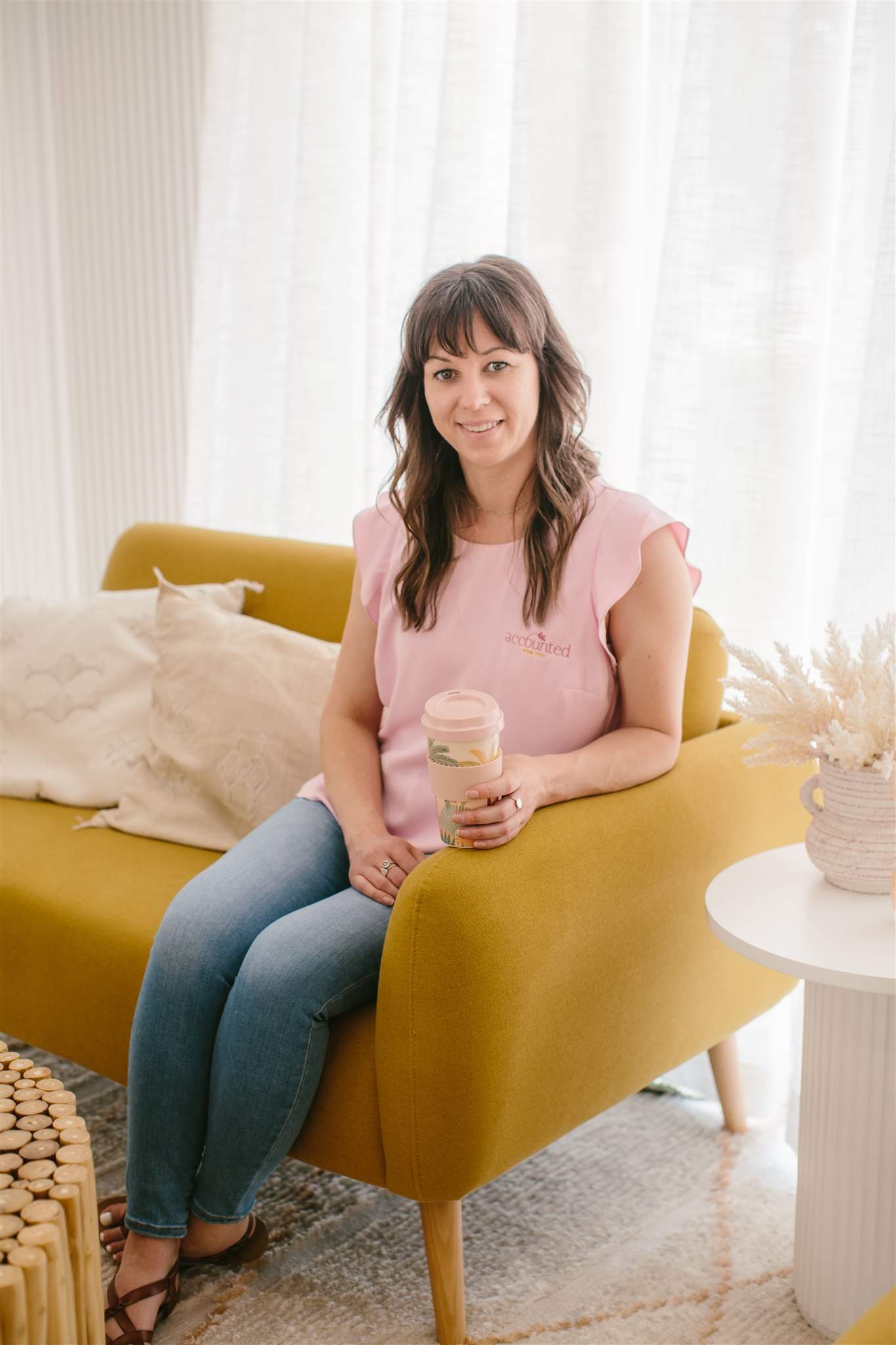 Rebecca Simon, a Bookkeeper from Accounted For You in a pink shirt sitting on a mustard couch, holding a cup of coffee.