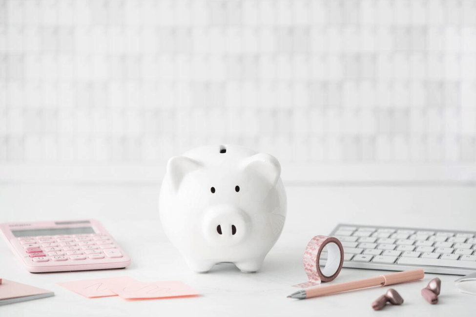 white piggy bank on a white table with pink calculator, sticky notes, washi tape, pen and apple keyboard