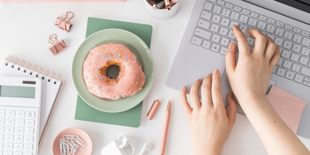 desktop flatlay with calculator, pink bulldog clips, pink donut on green plate and notebook, pink pen, hands typing on silver laptop.