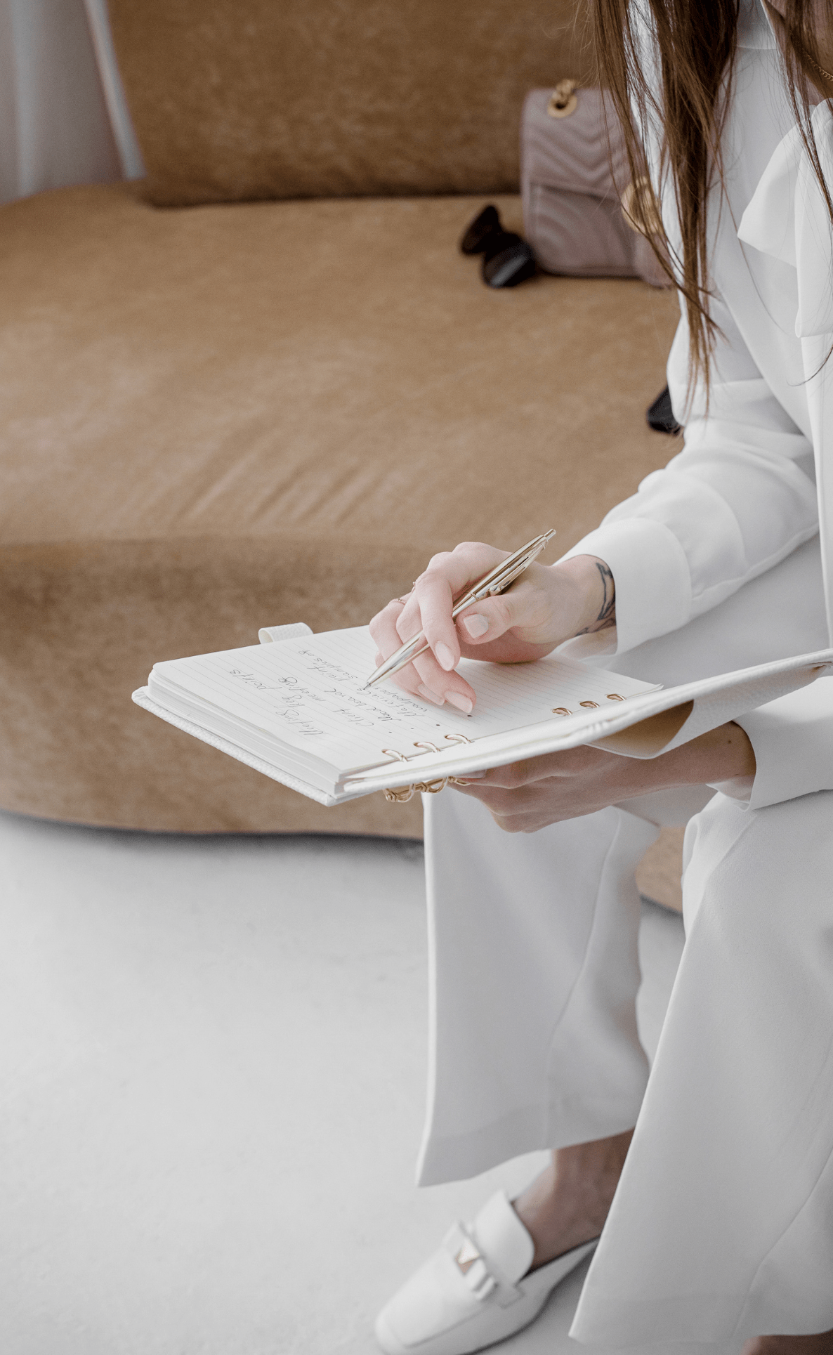women in white suit sitting on a tax couch with a pink handbag next to her, writing in a white planner