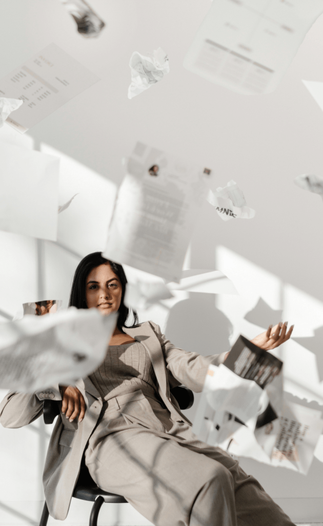 woman slouching back in chair wearing a grey business suit, throwing papers up in the air against a white background