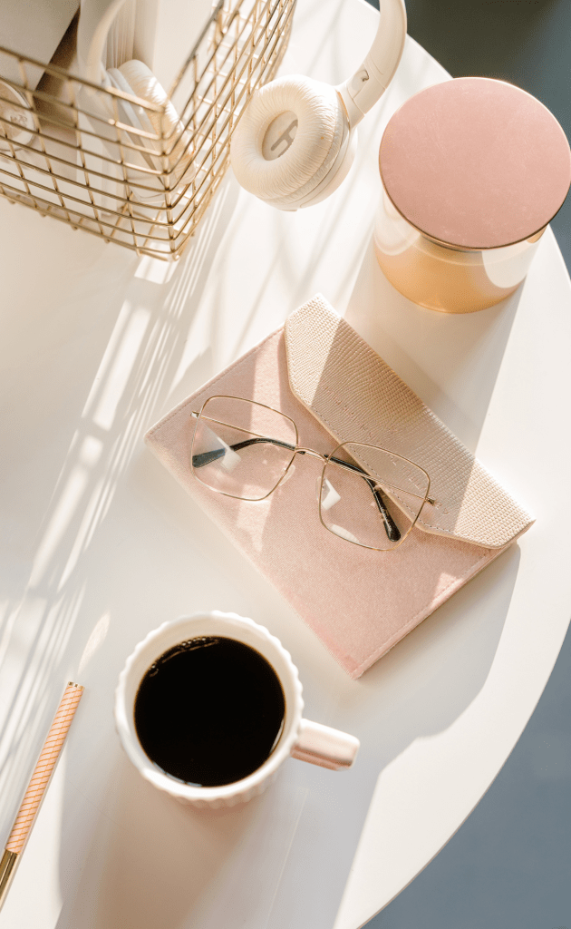 white table bathed in sunlight with pink notebook, reading glasses, cup of coffee, metal basket with white headphones, candle with rose gold lid and pink pen to side.