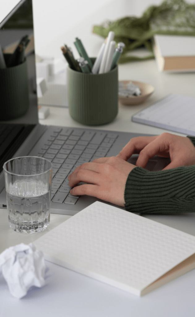 woman's hand in khaki sweater typing on chrome laptop with khaki pen pot, crumpled paper, notebook and glass of water to the side