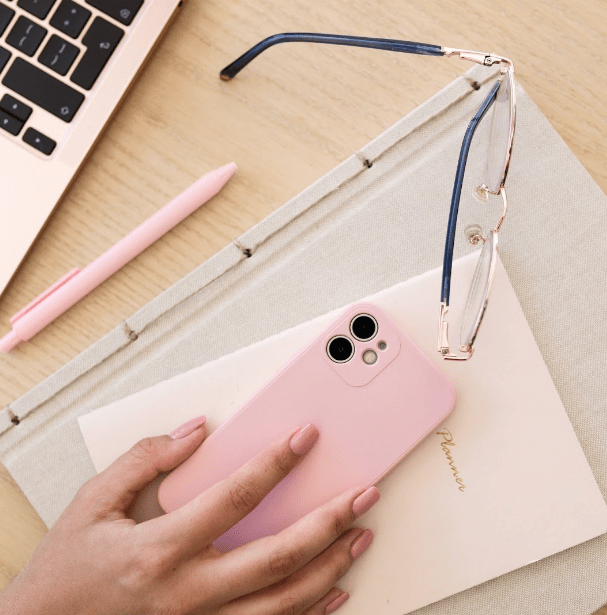 Hand holding pink phone with notebooks underneath. Laptop, pink pen and glasses to the side.