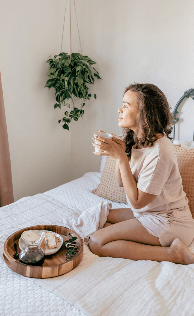 Girl sitting on bed in pink clothes holding a cup of coffee looking out the window with food platter in front of her on the bed
