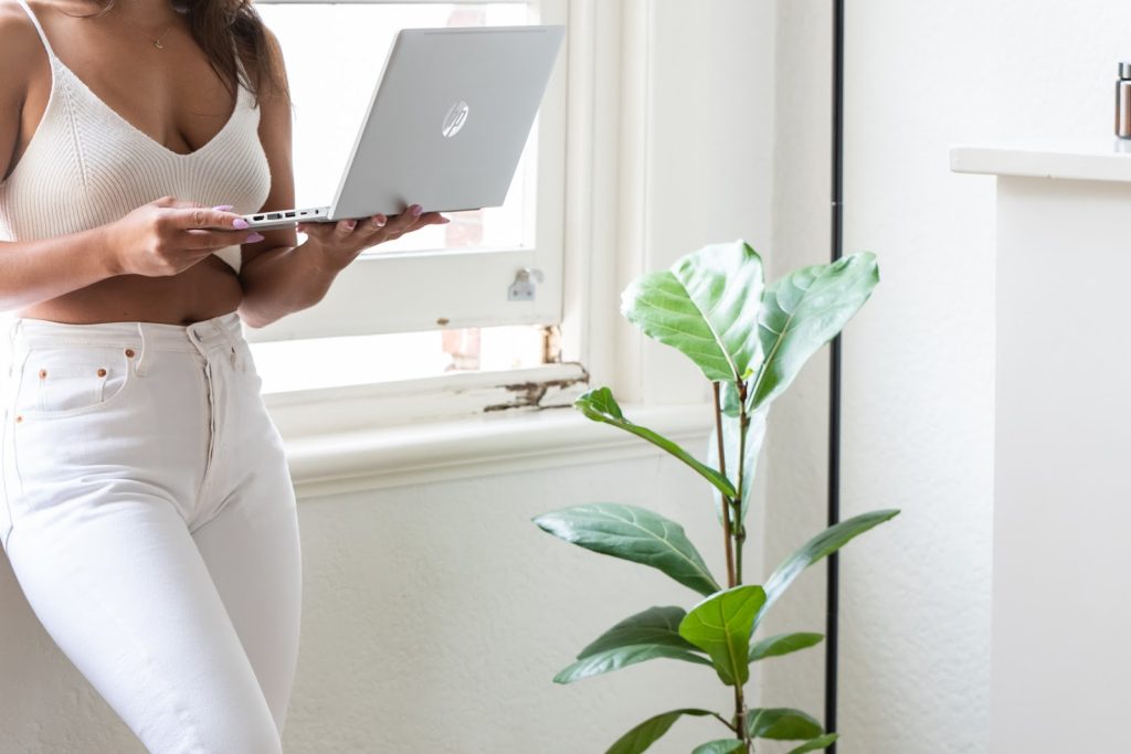 A woman in white jeans and singlet top leaning against an open window with a laptop in her hand. 