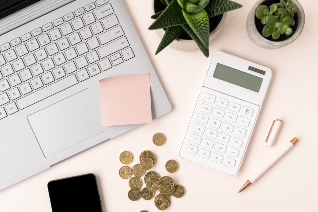 An open laptop, white calculator and Australian currency coins, laid on a pink desktop. 