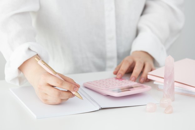person in a white shirt writing on a notepad with a pen using a calculator with pink crystals on the desk