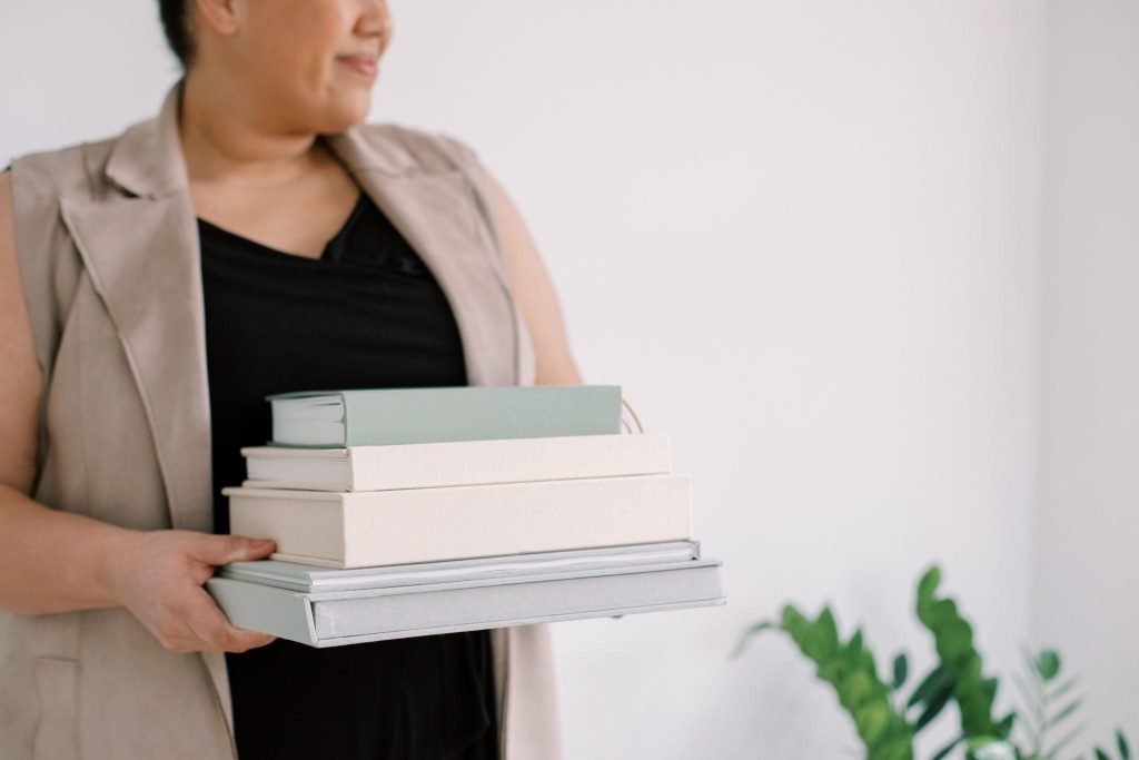 Female holding a stack of green and cream books against a white background, with a plant in the bottom corner. 