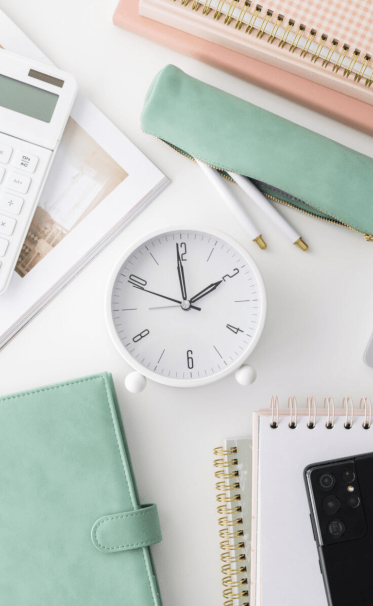 white flatlay with white clock in centre, green notebooks and pencil case surrounding