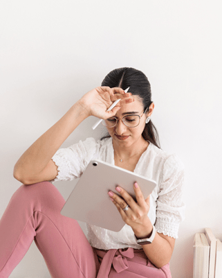 Woman in white top, pink pants leaning on a wall, hand on her forehead holding an ipencil, looking at her ipad