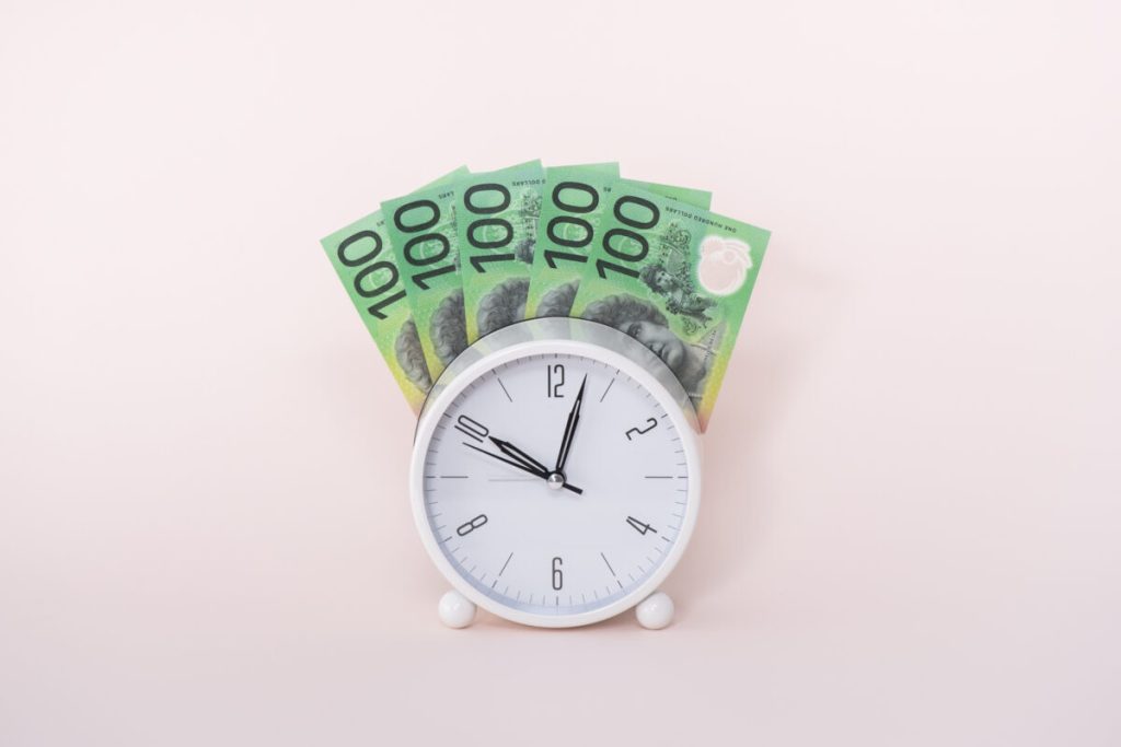 pink background. White round small clock, with green one hundred dollar notes fanned out behind it.