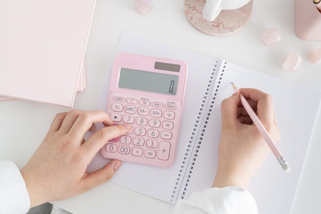 desk flatlay with person writing in white notepad, typing on pink calculator, with note pad and crystals scattered around.