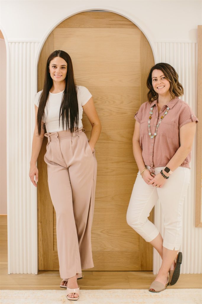 Rachel and Lisa, Xero bookkeepers from the Gold Coast leaning against a white and wood coloured wall. Both wearing pink and white tones.