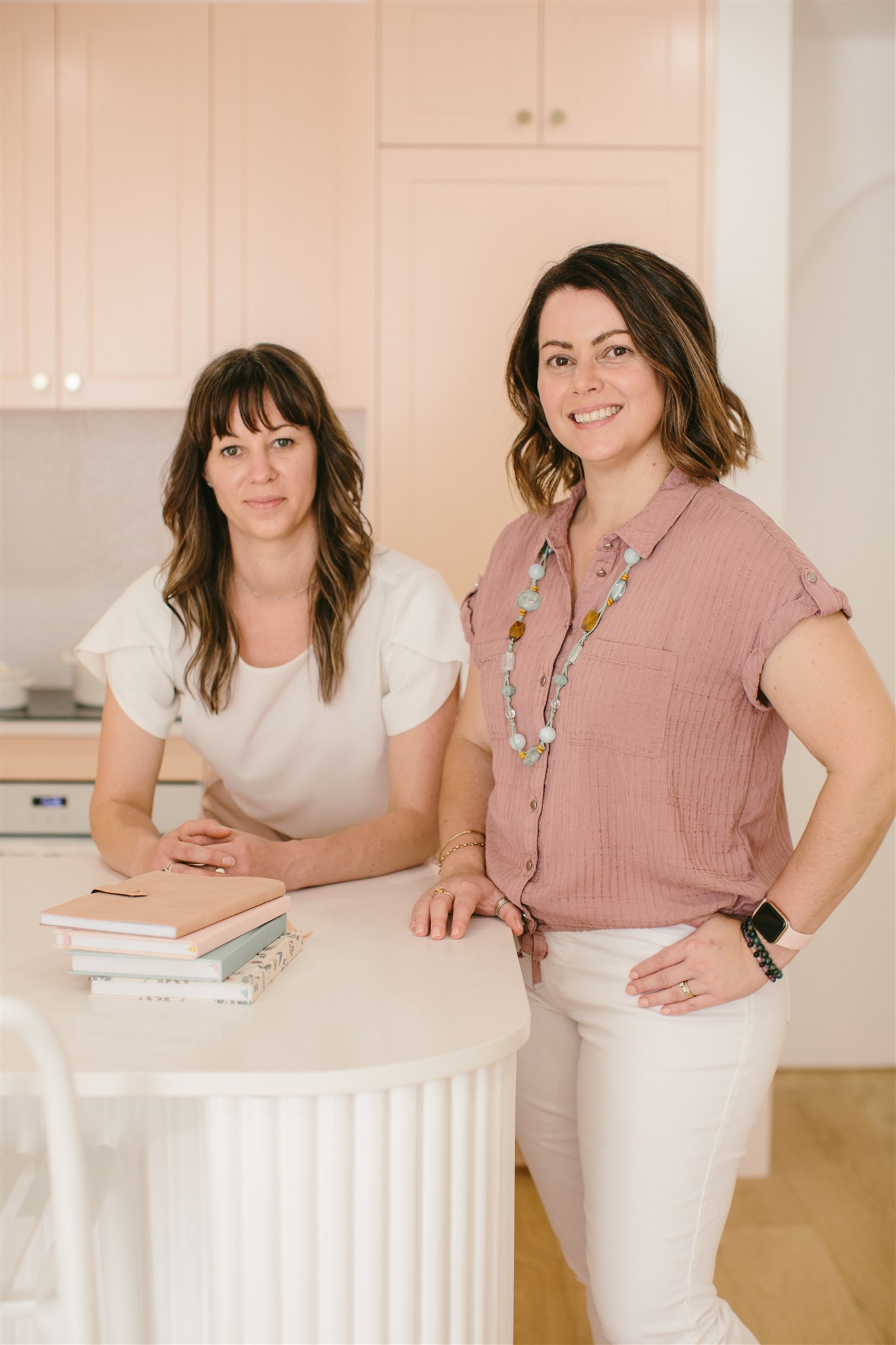 Rebecca and Lisa, Bookkeepers for women in business, resting on a white kitchen bench with a stack of notebooks in front. Both wearing pink and white tones.