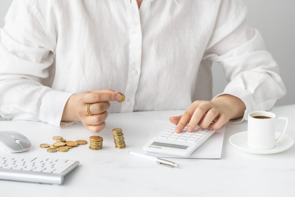 Woman sitting in a white shirt at a table counting gold coins and bookkeeping with a white calculator and white coffee cup. 