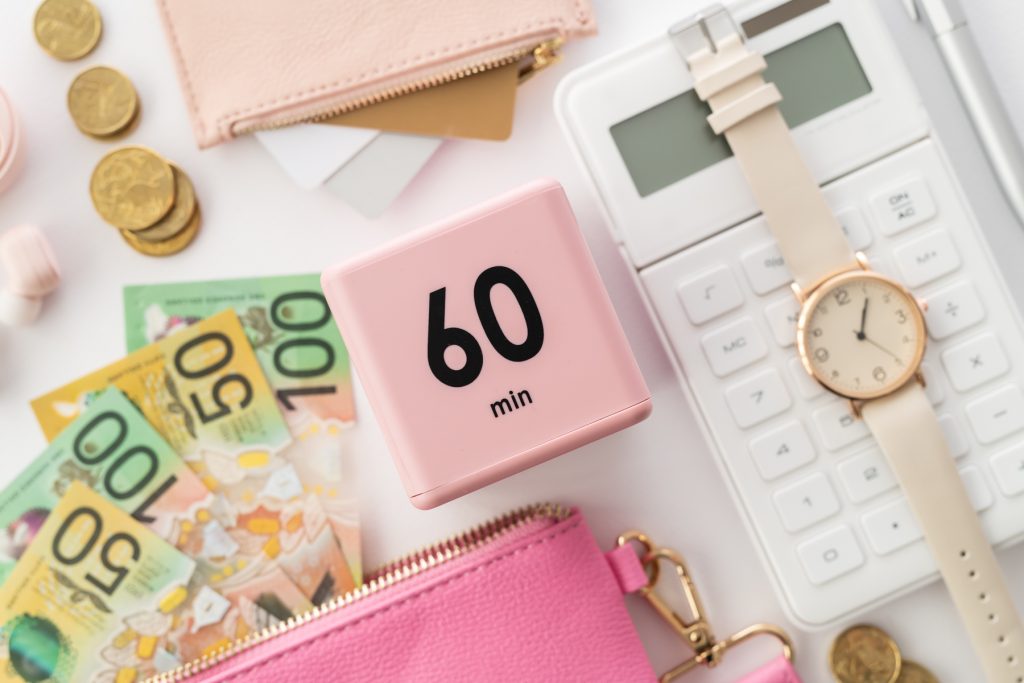 Flat lay image consisting of Australian currency in both notes and coins. A white calculator, a watch and pink coin wallets. 