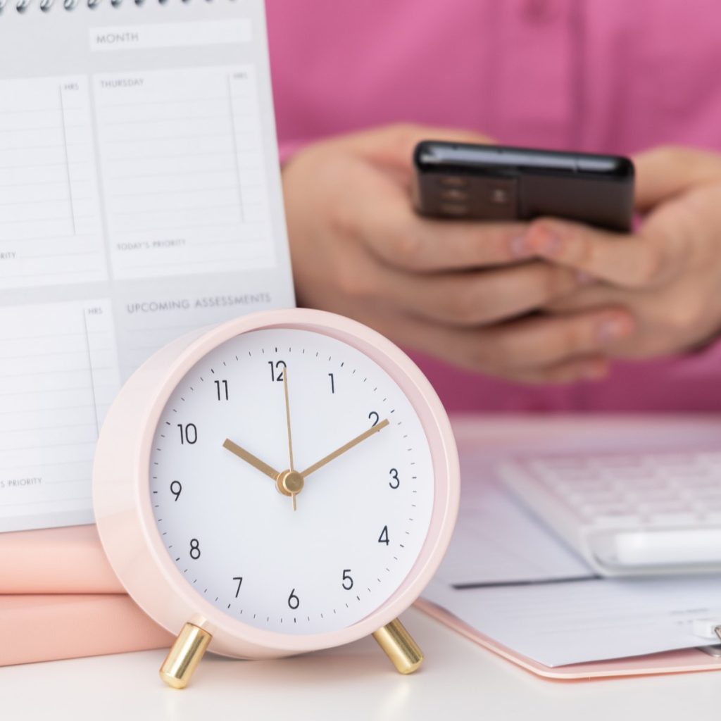 pink alarm clock in foreground with someone in pink shirt on their phone in the background, with notepads and calculator middle.