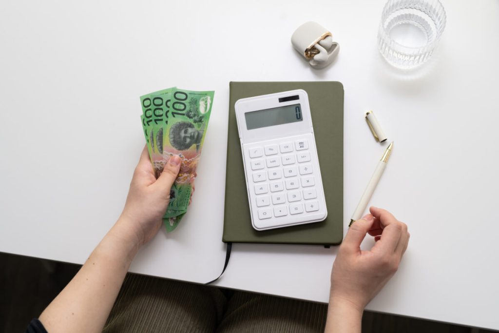 Overhead image of a person counting $100 Australian notes with a white calculator set upon an olive green notebook and a glass of water off to the side.