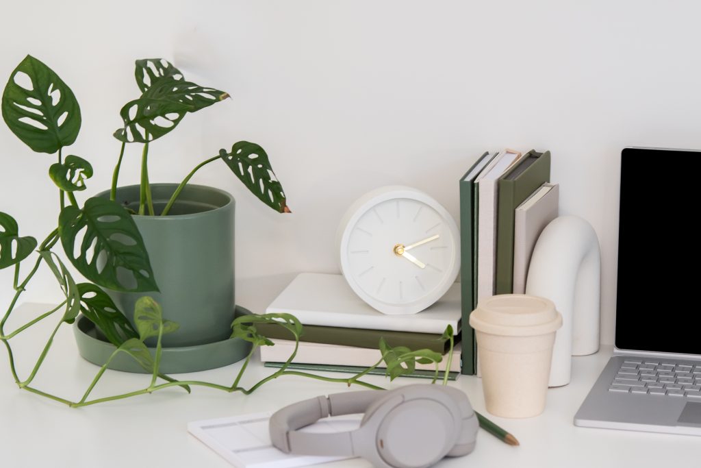 Work desk with a green plant in a pot, a pair of grey headphones, pink coffee cup and a laptop with a stack of green books beside it. 