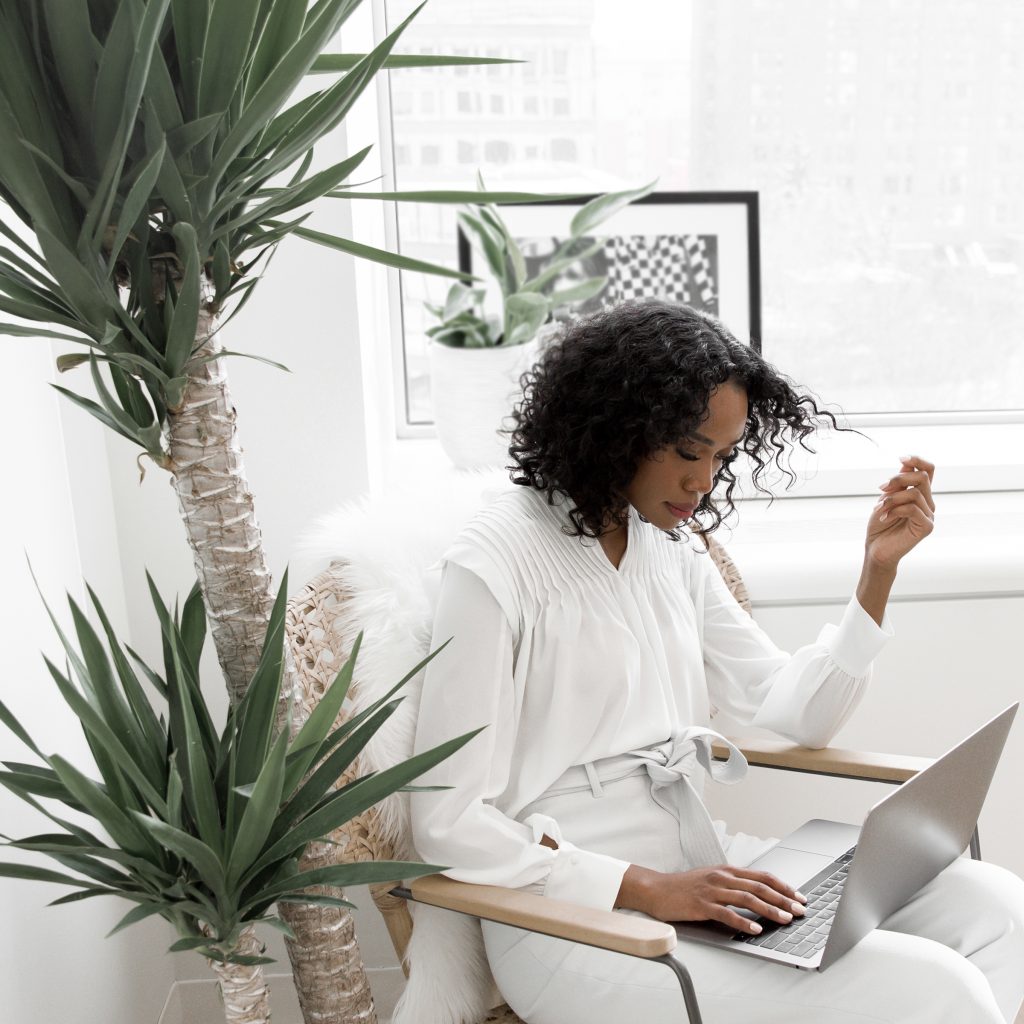 A woman dressed in white blouse and grey pants sitting on a chair looking at an open laptop.  Large green plant to her left side and a large window behind her. 