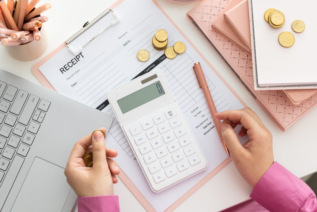 Persons hands holding a gold coin, overlooking a desktop with laptop, invoice clipboard, calculator and notepads.