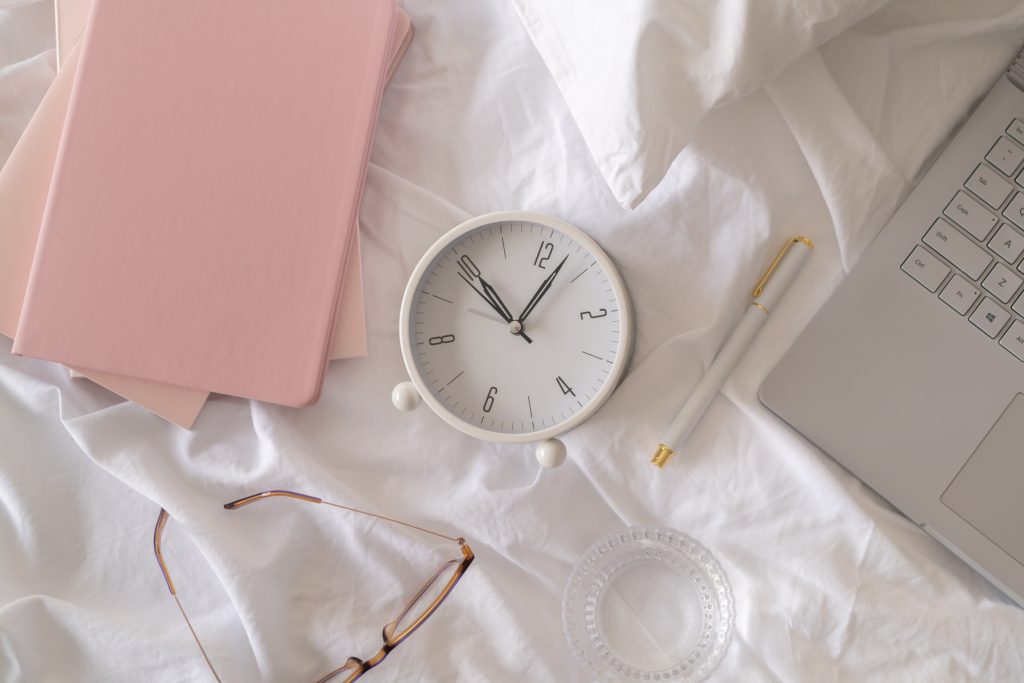 Laptop, white clock, pen, and pink notepads on a white linen sheet.