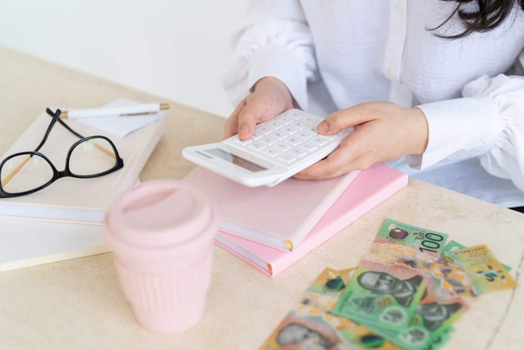 peachy marble desk, with a pink travel coffee cup in the foreground, some $100 and $50 notes on the side, someone typing on a white calculator stacked on some notebooks.