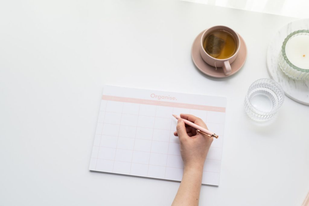 white desk, with someone writing on a white organise notepad with a cup of tea, cup of water and candle to the side.