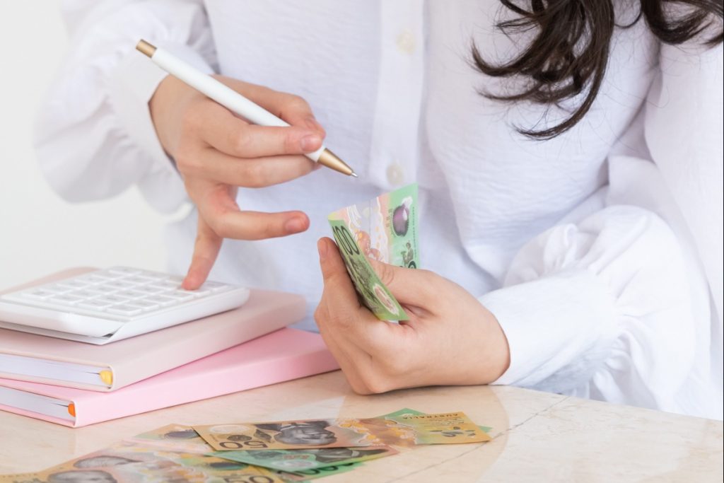 someone with long brown hair, in a white linen shirt, counting $100 & $50 notes, while using a white calculator stacked on top of pink notebooks.