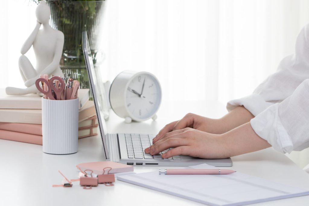 someones arms in a white linen shirt, typing on a silver laptop on a white desk side cropped view, with a stack of pink notebooks behind, white container of pink pens and other stationery, with a white clock in the background.