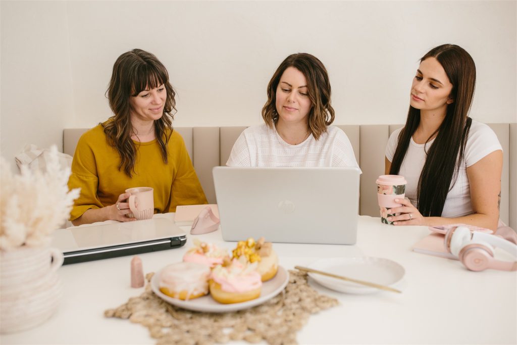 Rebecca, Lisa & Rach on a neutral bench seat, at a round table, looking at a silver laptop intently. With other desk items in the foreground, including donuts!