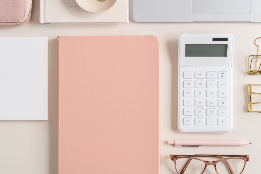 Peachy pink flatlay desk, with peach notepad, pink pen, white calculator, gold clips and other mixed stationery.